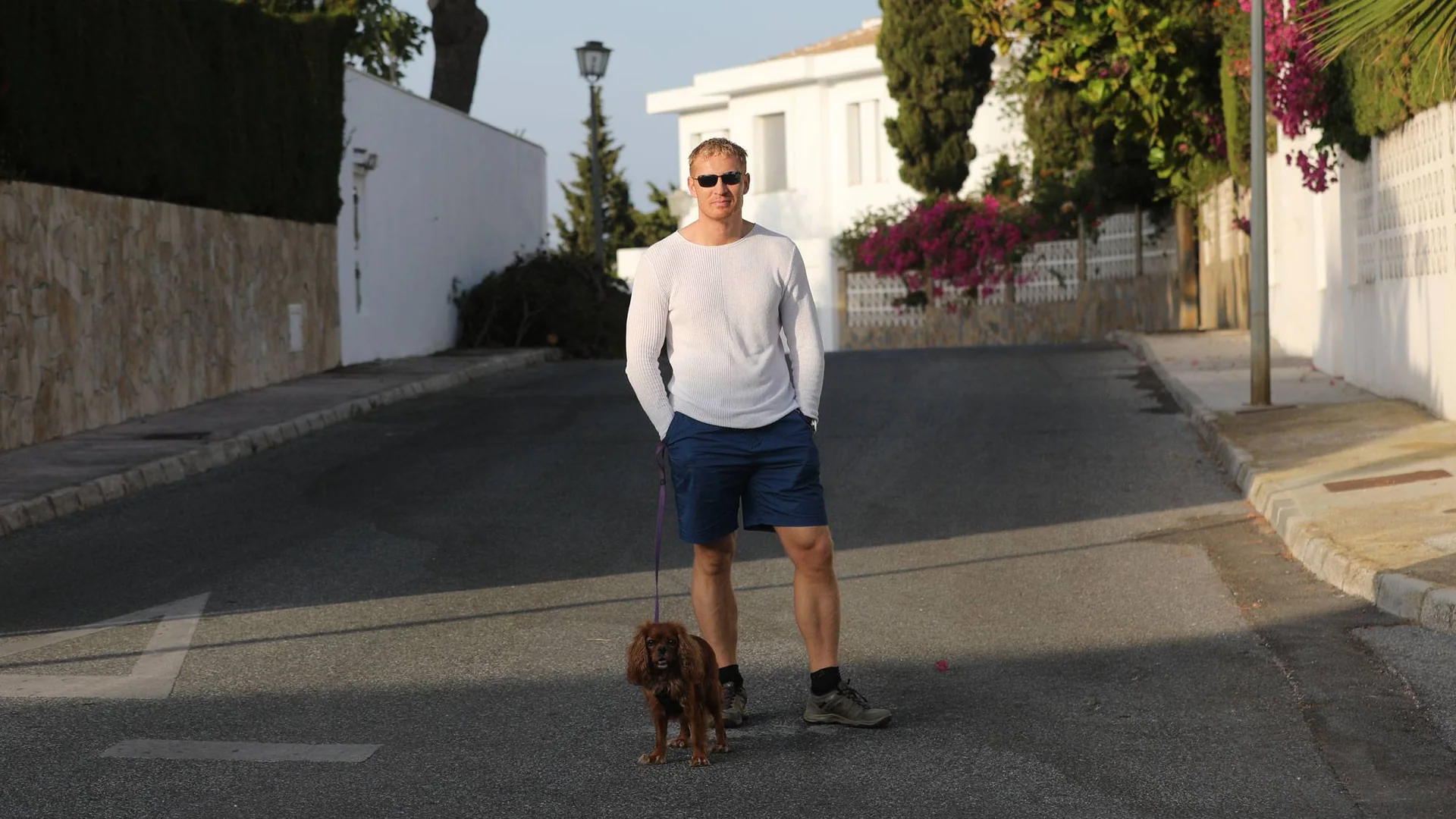 En mann med en liten hund står i en gate med hvite murer på sidene.