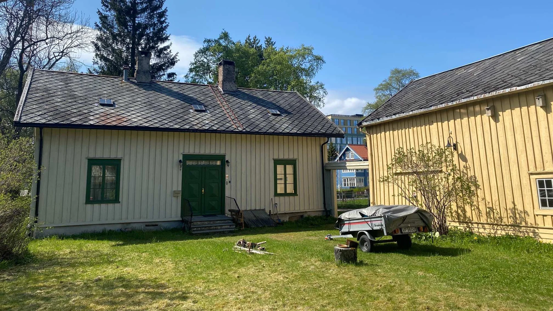 Fronten av et gammelt hvitt hus med grønn dør og grønne lister. På plenen foran står en tilhenger, og i siden av bildet ser man litt av en gul låve.