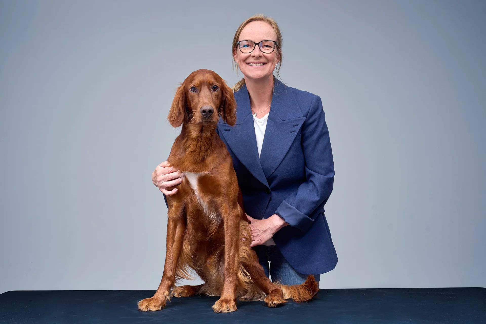 Smiling woman in a blue blazer posing with a brown Irish Setter in a studio backdrop of gray walls and a dark table