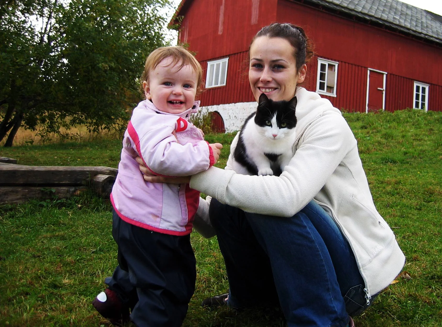 En kvinne og et barn holder en svart og hvit katt foran en rød bygning.