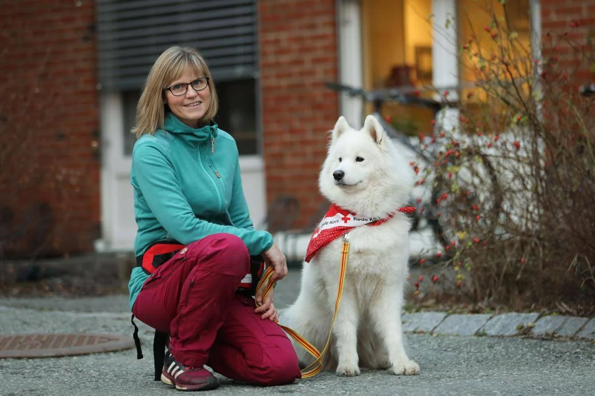 En kvinne sitter på huk ved en hvit hund med rødt tørkle utenfor et hus.