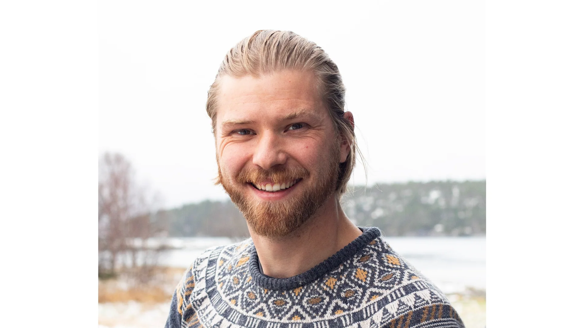 Close-up portrait of a smiling man with blond hair and a beard, wearing a patterned knit sweater outdoors in winter.