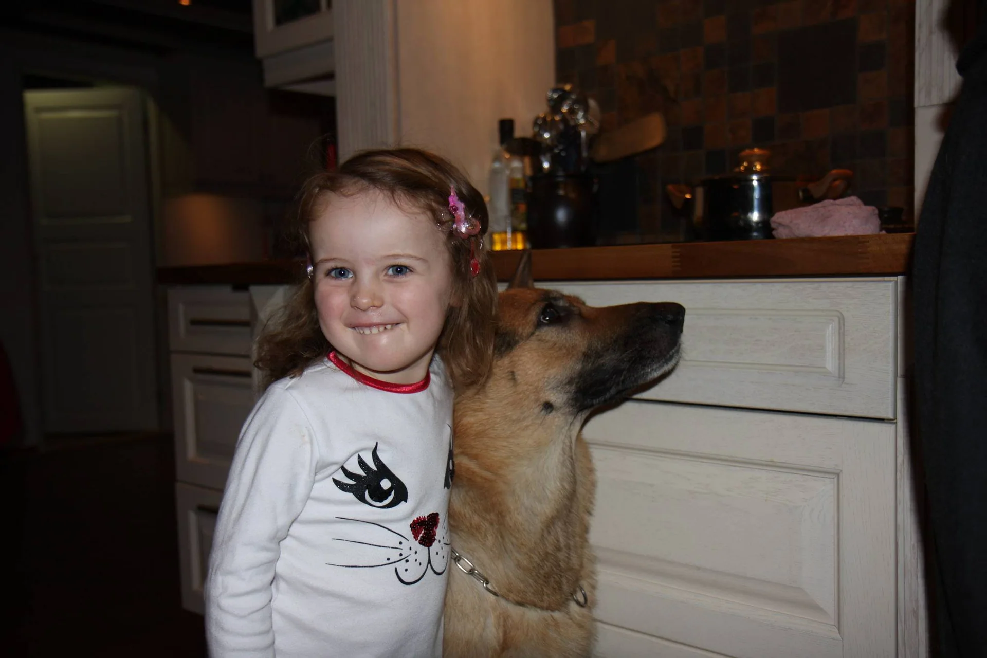 Et barn står tett inntil en hund på et kjøkken.