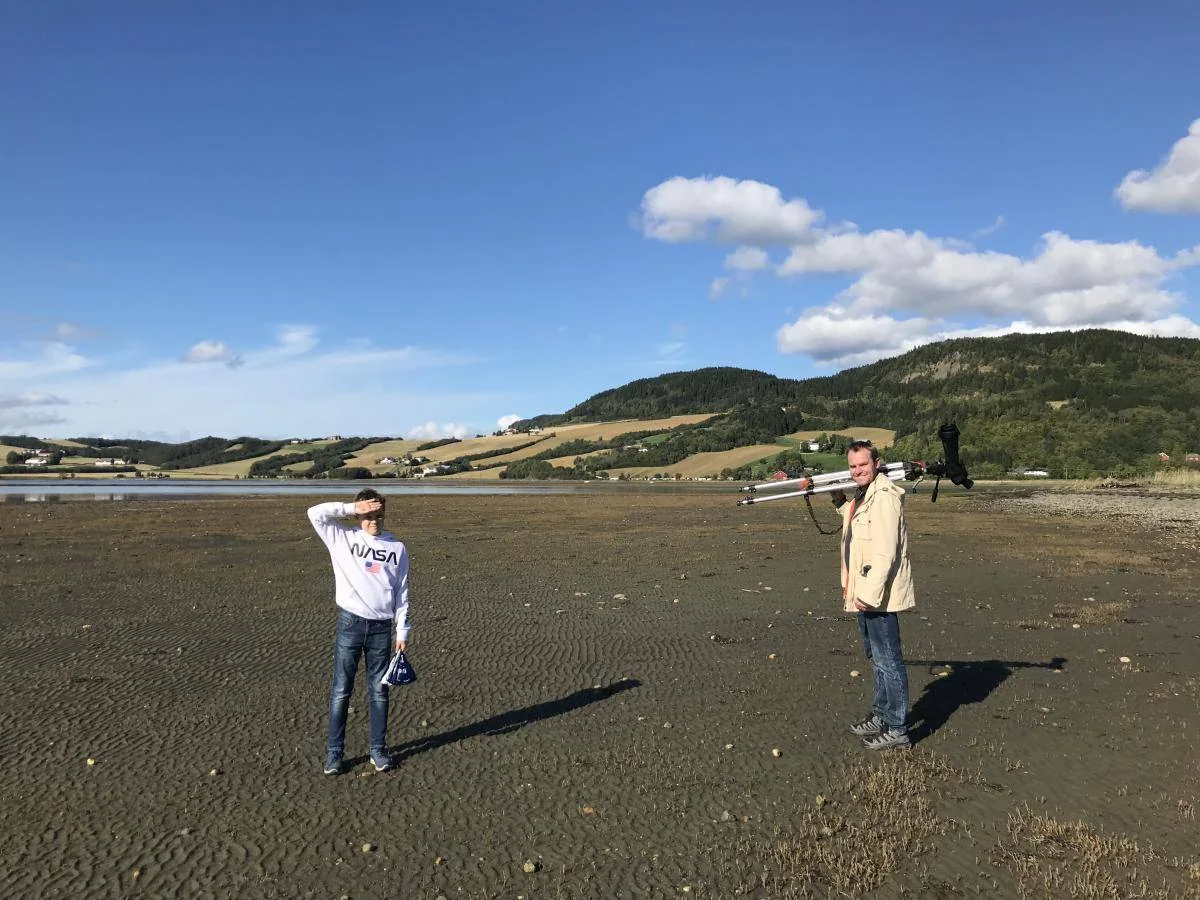 To personer står ute på en fjære med sanddyner og åser i bakgrunnen.