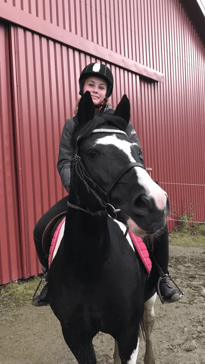 En rytter med hjelm sitter på en svart hest ved en rød låve.