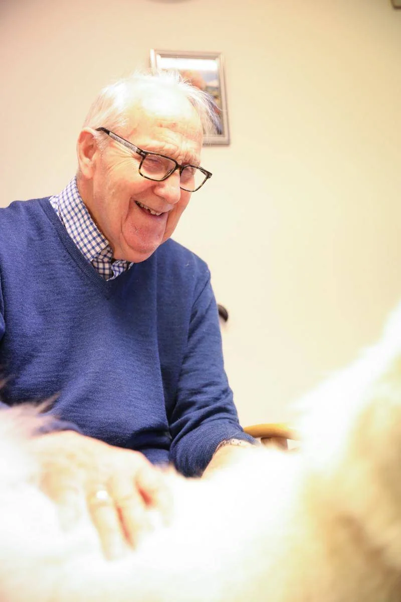 En smilende mann klapper en hvit hund ved stolen sin.