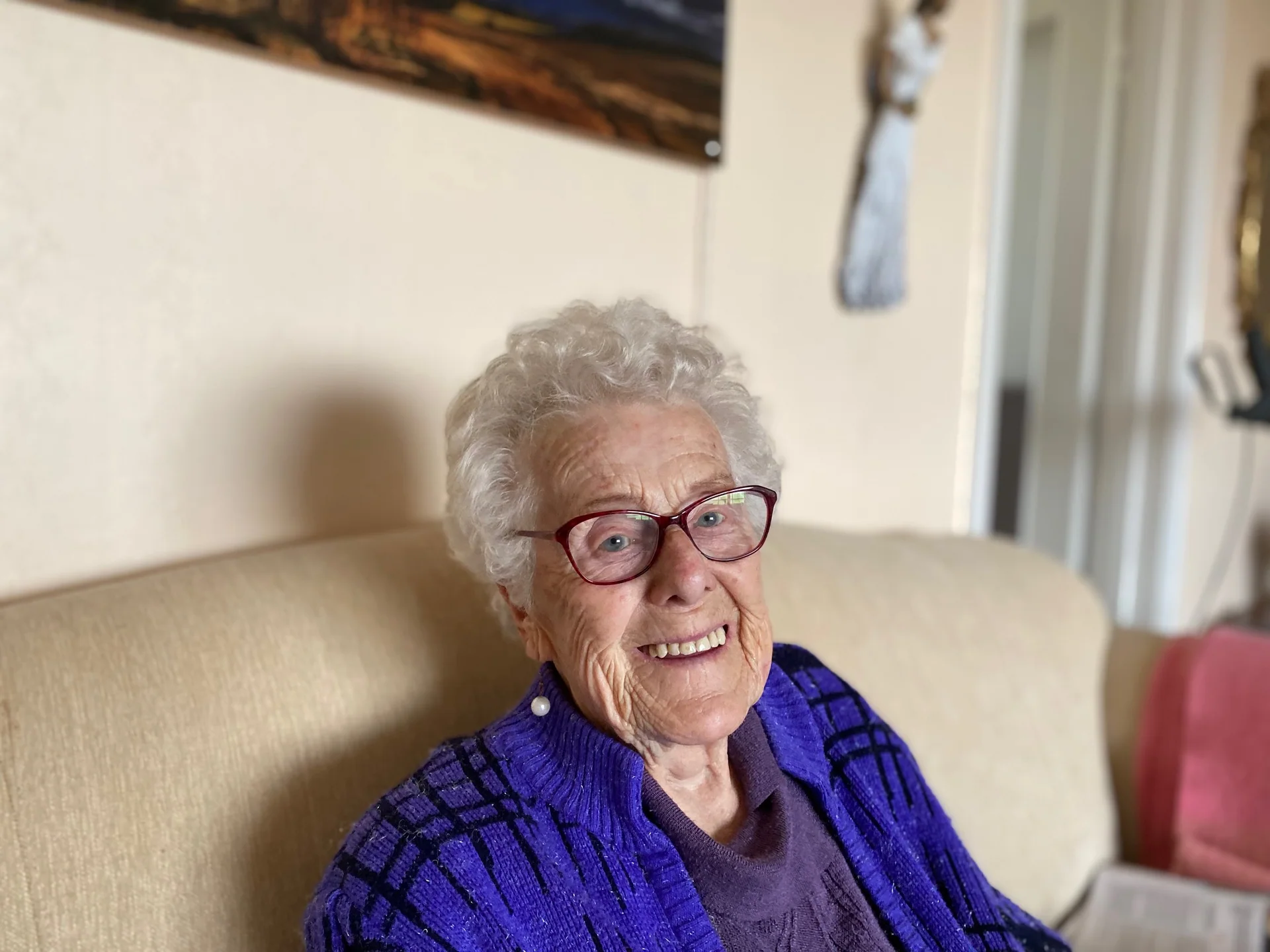 En smilende kvinne med briller og lilla jakke sitter i en sofa.