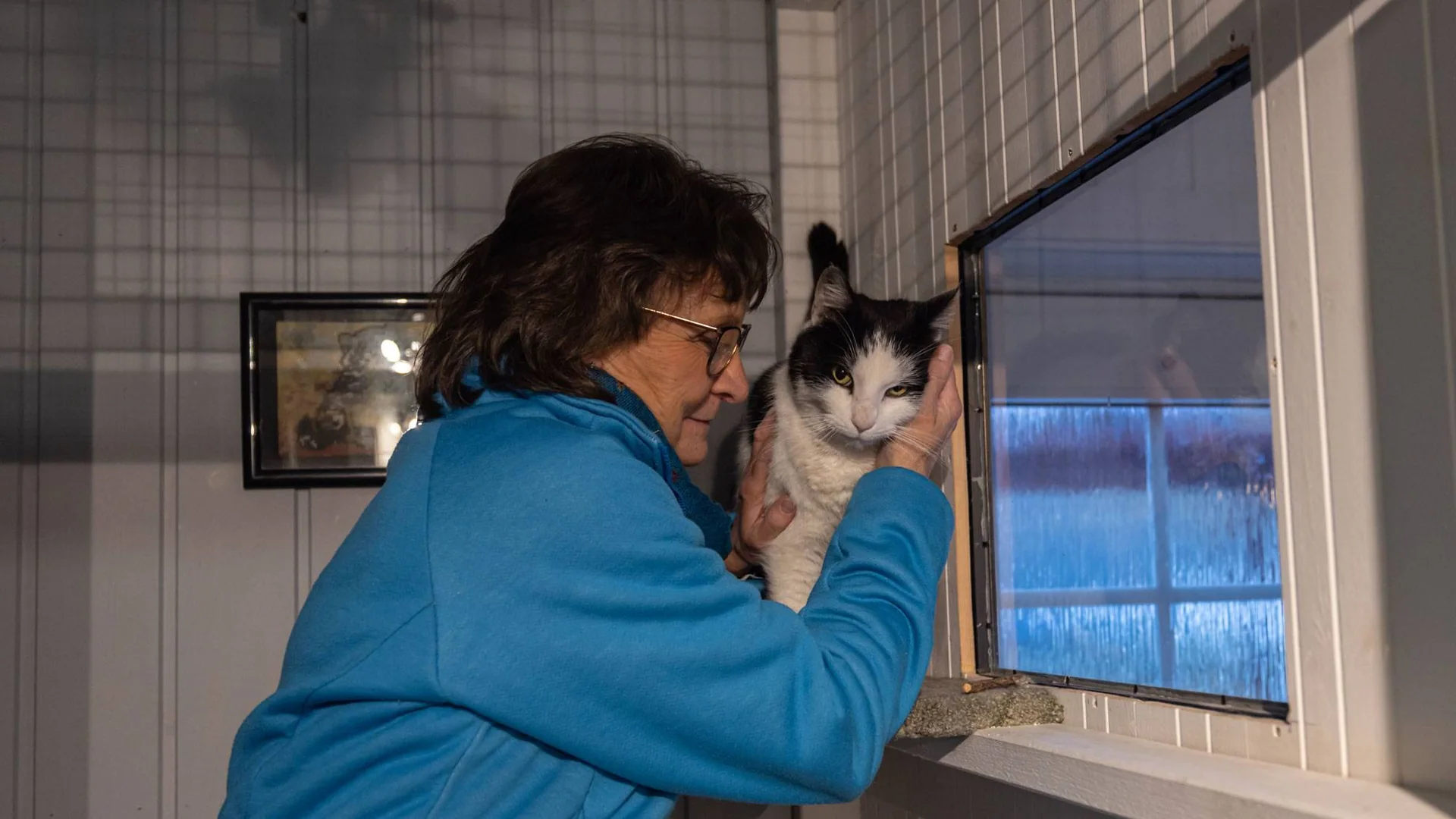En kvinne holder en svart og hvit katt inntil ansiktet ved et vindu.