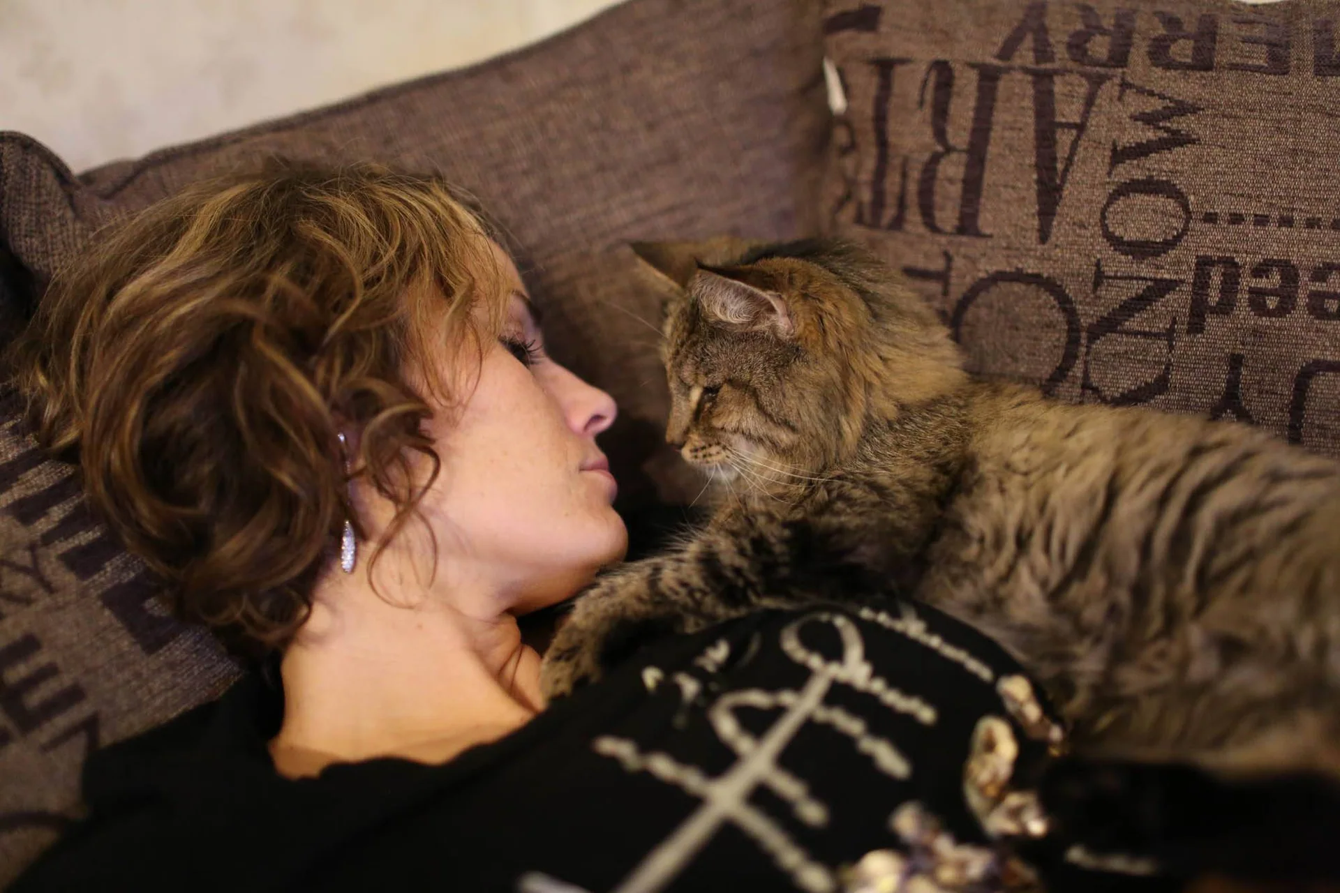 En kvinne ligger i sofaen med en stripete katt tett inntil ansiktet.