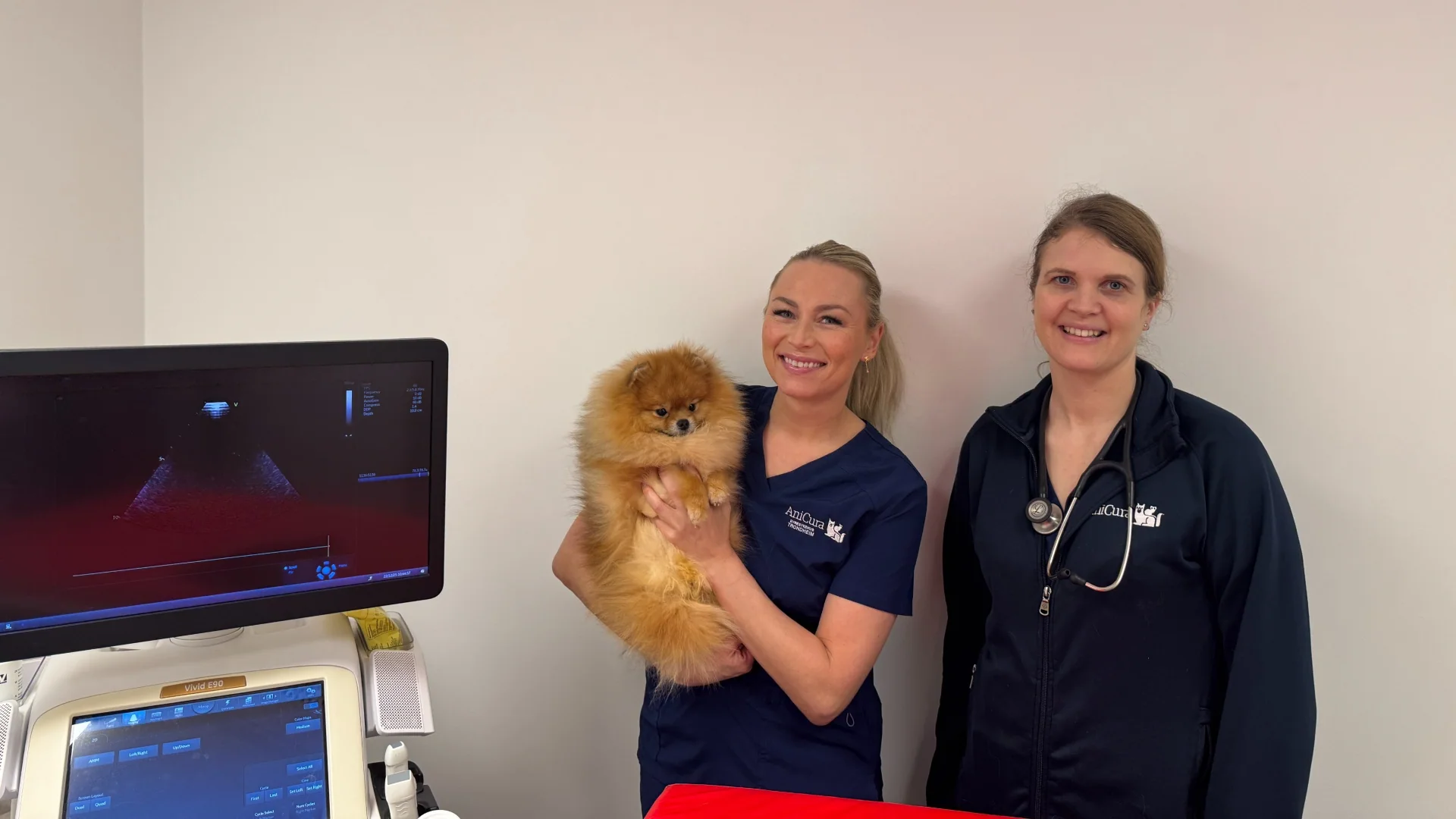 Camilla Kristoffersen og Ine Sund står ved siden av en ultralysmaskin på dyreklinikken. Kristoffersen holder en pomeranier i armene.