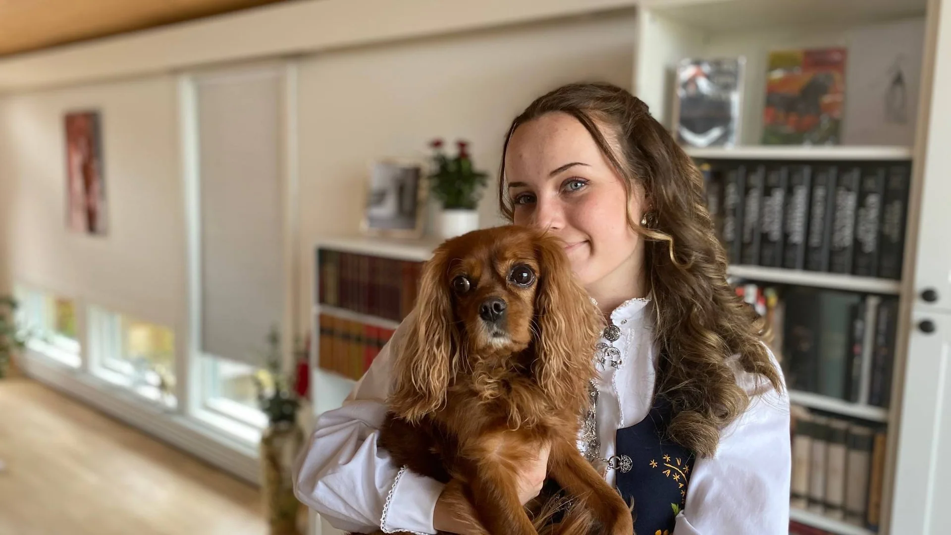 Ung jente i bunad holder en hund. Bokhyller i bakgrunnen.