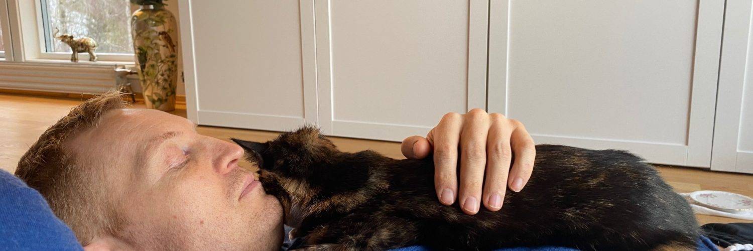 Mann i blå genser ligger på gulvet med en mørk katt på brystet.