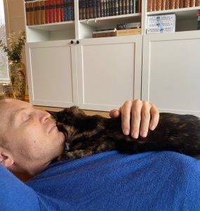 Mann i blå genser ligger på gulvet med en mørk katt på brystet.