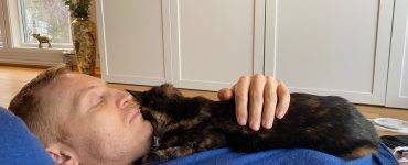 Mann i blå genser ligger på gulvet med en mørk katt på brystet.