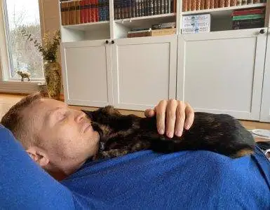 Mann i blå genser ligger på gulvet med en mørk katt på brystet.