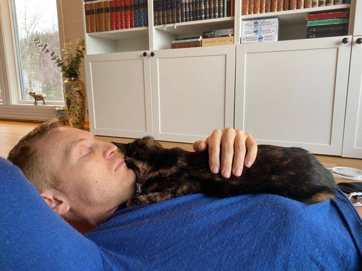 Mann i blå genser ligger på gulvet med en mørk katt på brystet.