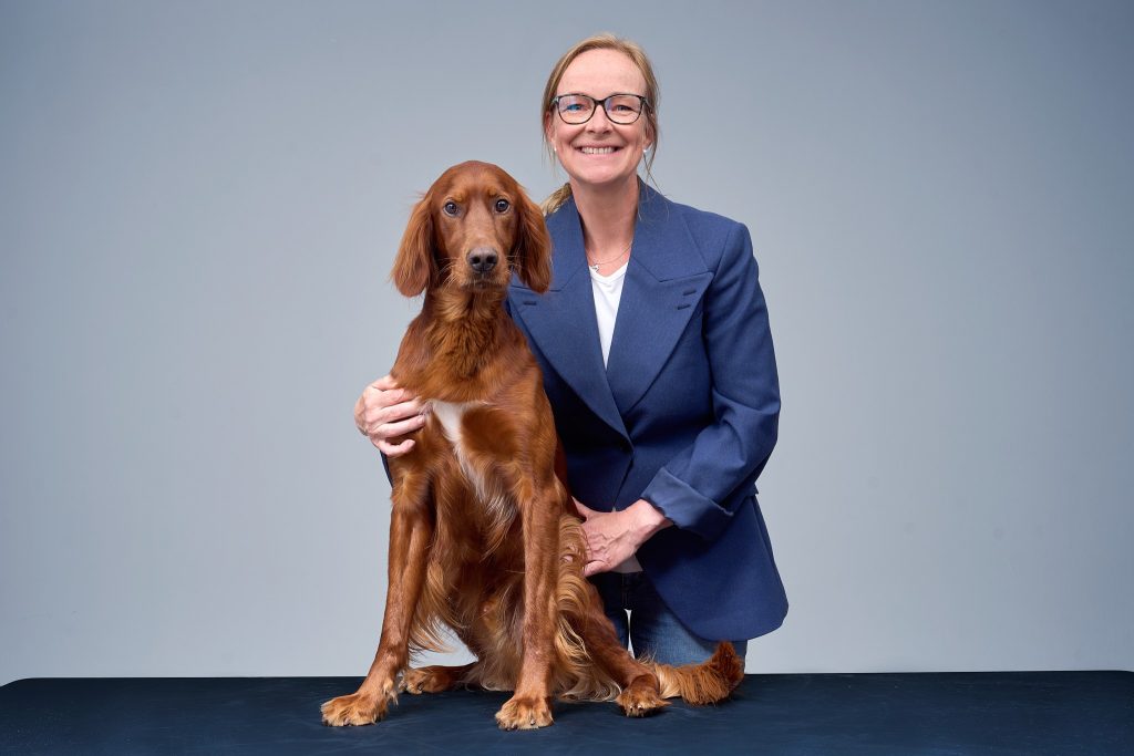 Hanne Christine Øverli 4230 WEB - Dyr og folk Smiling woman in a blue blazer posing with a brown Irish Setter in a studio backdrop of gray walls and a dark table