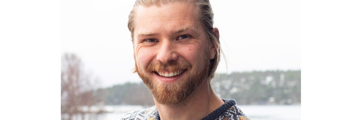 Close-up portrait of a smiling man with blond hair and a beard, wearing a patterned knit sweater outdoors in winter.