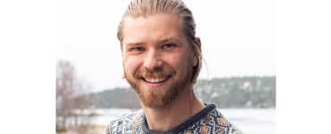 Close-up portrait of a smiling man with blond hair and a beard, wearing a patterned knit sweater outdoors in winter.