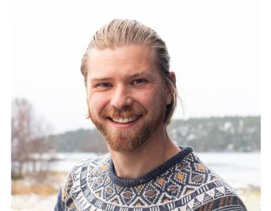 Linas Johan Hauge Wenghaug - Dyr og folk Close-up portrait of a smiling man with blond hair and a beard, wearing a patterned knit sweater outdoors in winter.